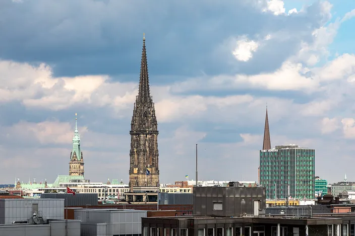 The blackened stone spire of the ruined St. Nikolai church rising above the flat modern rooftops of central Hamburg, with the green copper tower of the town hall to the left and a slender brown church spire beside a green glass office block to the right, under a sky of broken cloud.