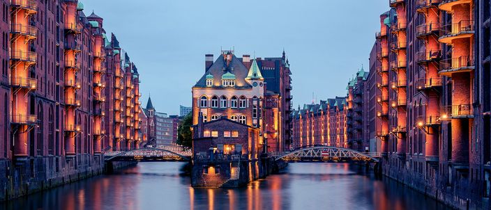 A red-brick canal in Hamburg's Speicherstadt at blue hour, warehouse blocks with iron balconies lit warm orange on both banks and converging on the Wasserschloss — a small turreted brick building marooned on its own island, with a slate roof, copper dormers and a clock turret — flanked by two white iron footbridges, the whole scene mirrored in still dark water.