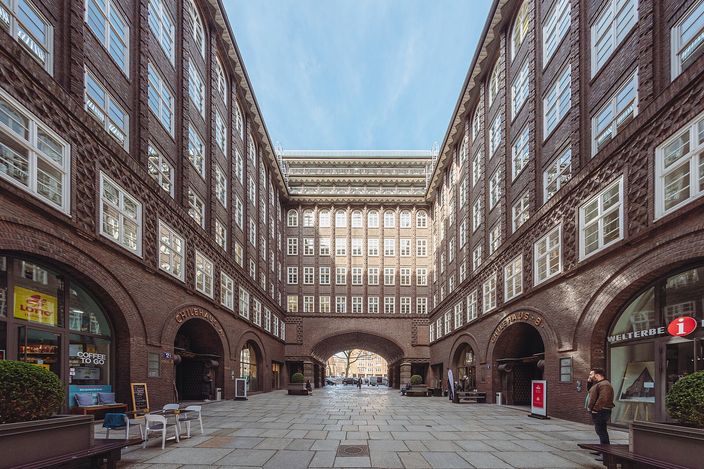 The arcaded inner courtyard of the Chilehaus in Hamburg: dark brown brick office storeys with white-framed windows rising on three sides above a ground-floor colonnade of round brick arches, an archway at the far end opening through to the street, the paving empty apart from a few passers-by.