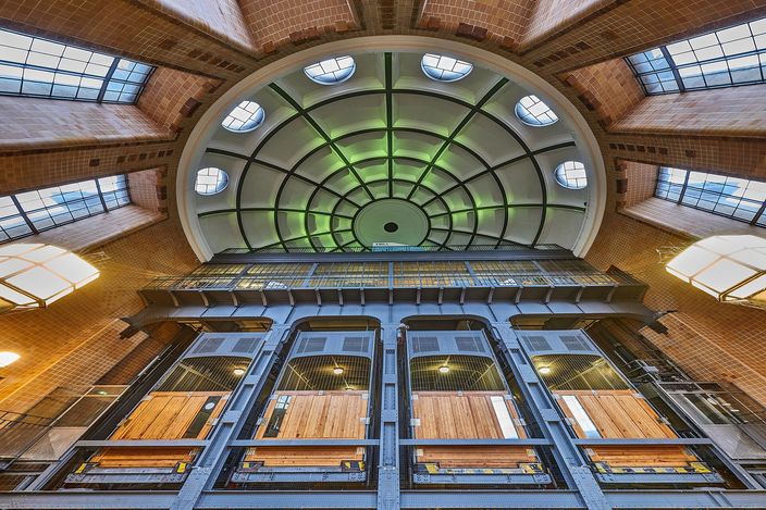 The domed entrance hall of Hamburg's old Elbe tunnel seen from below: a green glazed cupola on radiating iron ribs above tiled brick walls and clerestory windows, with four tall riveted-steel lift shafts standing side by side beneath it, their wooden-floored cars visible through wired glass.