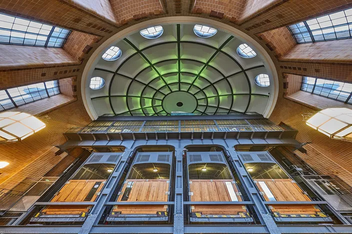 The domed entrance hall of Hamburg's old Elbe tunnel seen from below: a green glazed cupola on radiating iron ribs above tiled brick walls and clerestory windows, with four tall riveted-steel elevator shafts standing side by side beneath it, their wooden-floored cars visible through wired glass.