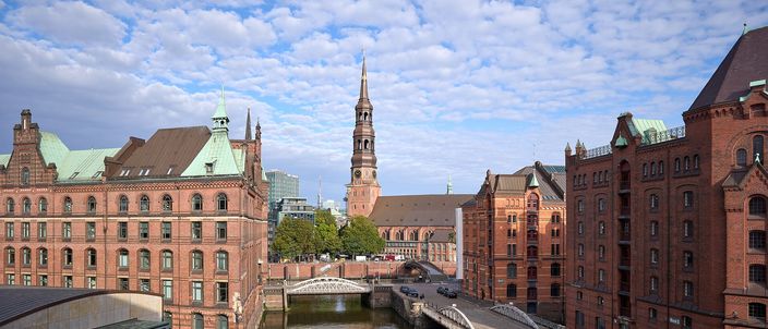 A canal in Hamburg's Speicherstadt seen from a bridge: seven-storey red-brick warehouses with green copper gables and iron loading balconies lining both banks, a green arched bridge and a white iron footbridge crossing the water, and the dark spire of St. Katharinen closing the view under broken cloud.