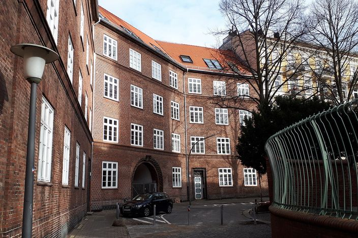 A red-brick residential courtyard block at St. Georgs Kirchhof in Hamburg-St. Georg, four storeys with white-framed windows and a red-tiled roof, a bare tree and modern lamppost in the foreground on a clear winter day.