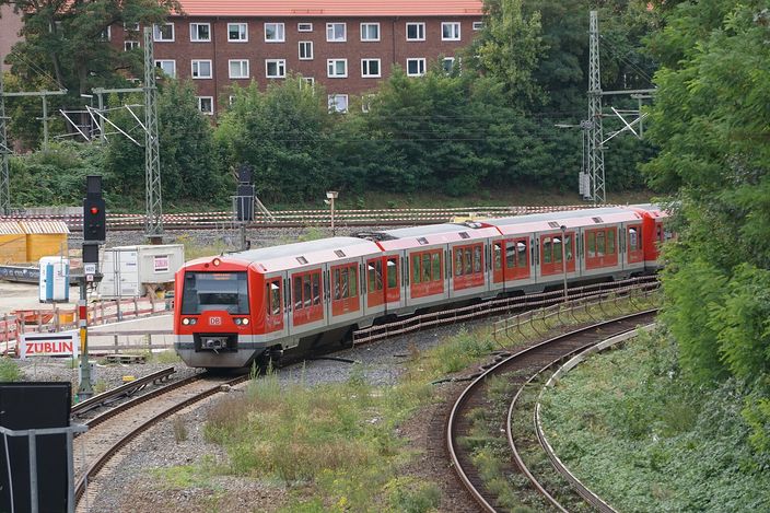 A red Hamburg S-Bahn train (Class 474) curving through a cutting past red-brick apartment blocks on the S1 line