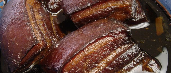 Close-up of thick, glossy dark reddish-brown braised pork belly squares with the skin on top, showing alternating bands of fat and lean meat sitting in a rich reduction.