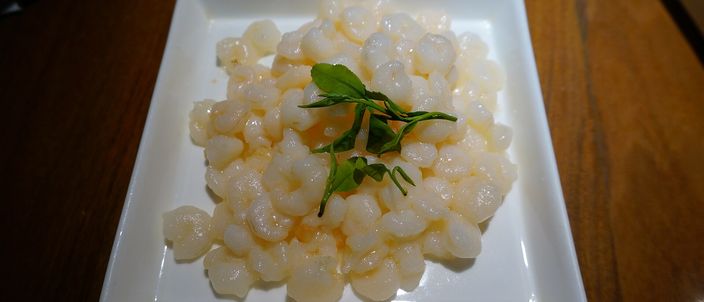 A small square white plate of pale, ivory shrimp, barely curled, studded with a few whole green tea leaves, on a dark wood restaurant table.