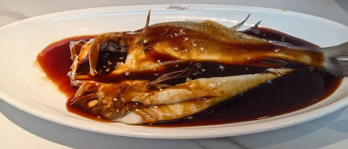 A whole vinegar-glazed grass carp, split lengthwise, plated on an oval white dish with a thin, glossy reddish-brown sauce pooling around the fish rather than coating it thickly.