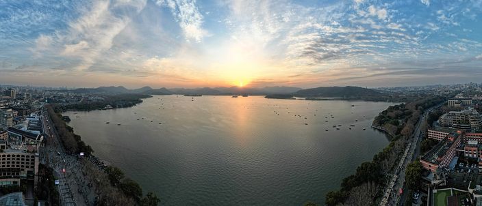 Aerial panorama of West Lake at sunset, a causeway crossing the water, small boats scattered on the lake, and hills and the city skyline framing the shore.