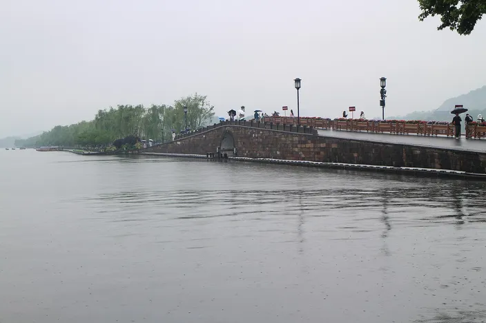 Stone Broken Bridge on West Lake's Bai Causeway under a hazy sky, with pedestrians and lanterns lining the balustrade and willows along the shore.