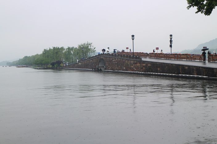 Stone Broken Bridge on West Lake's Bai Causeway under a hazy sky, with pedestrians and lanterns lining the balustrade and willows along the shore.