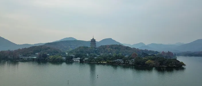 West Lake seen across open water toward a wooded shoreline, the multi-tiered Leifeng Pagoda rising above the trees under a hazy pale sky.