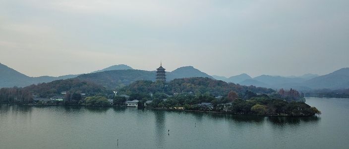 West Lake seen across open water toward a wooded shoreline, the multi-tiered Leifeng Pagoda rising above the trees under a hazy pale sky.