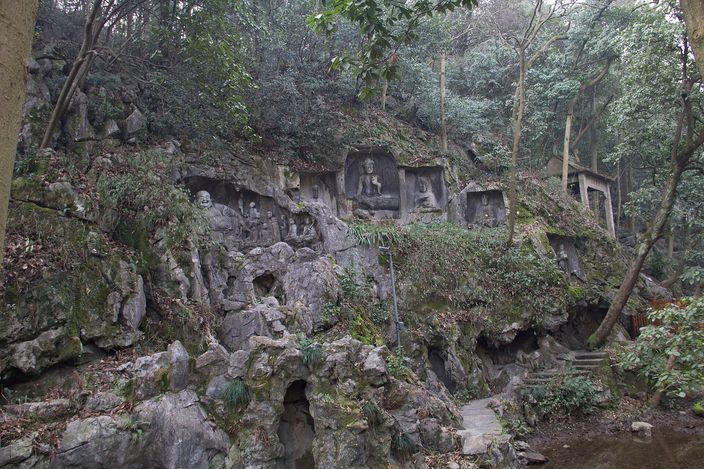 Buddhist figures carved directly into a moss-covered limestone cliff face at Feilai Feng, half-hidden among trees.