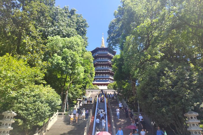 Crowds climbing stone steps up to Leifeng Pagoda through wooded parkland on West Lake's shore, on a sunny day.