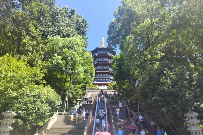 Crowds climbing stone steps up to Leifeng Pagoda through wooded parkland on West Lake's shore, on a sunny day.