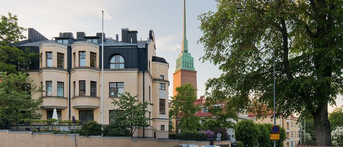 A curved cream-coloured Jugend apartment house on a quiet Eira street, cars parked along the kerb, with the green copper spire of Mikael Agricola Church rising behind and a large leafy tree framing the right side, in summer evening light.