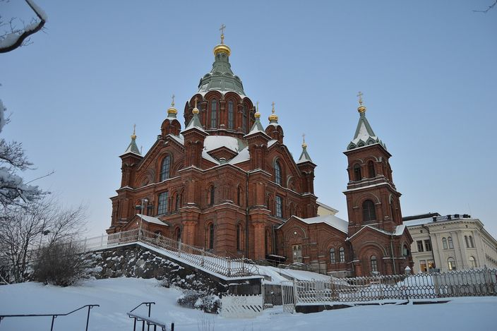 A red-brick cathedral with green copper roofs and gold onion domes standing on a snow-covered rocky rise, a granite staircase with white railings climbing to its door under a pale winter sky.