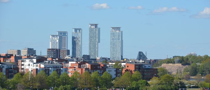 A cluster of tall glass and concrete towers rising behind low red-brick apartment blocks on a green wooded shoreline, seen across open blue water with a small yellow water bus crossing in the foreground.