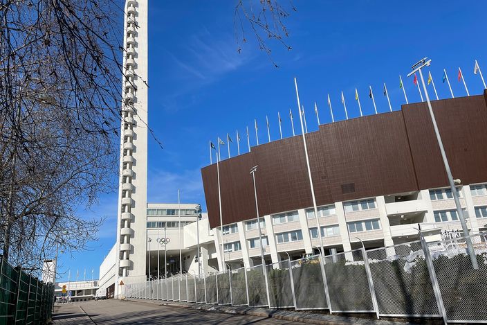 The white functionalist tower of the Helsinki Olympic Stadium against a deep blue sky, with the stadium's brown-clad stand, a long row of flagpoles and the Olympic rings below it, seen from the street on a bright early-spring day.