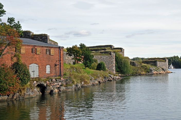 A red-brick building and grass-topped earth ramparts over granite revetments running along the water's edge of the Suomenlinna sea fortress, with a small sailing boat out on the strait beyond.