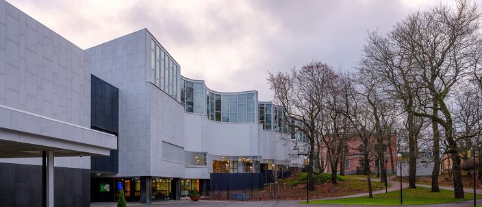 The white marble-clad flank of Finlandia Hall under an overcast sky, its angular glazed upper volume rising above a long low canopy, with bare trees and an empty granite forecourt in front.