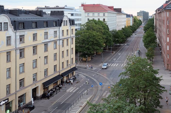 Elevated view of Helsinginkatu in Kallio: a curving tram-tracked avenue lined with cafés under a yellow National Romantic apartment block, tree-lined median and taller blocks receding north