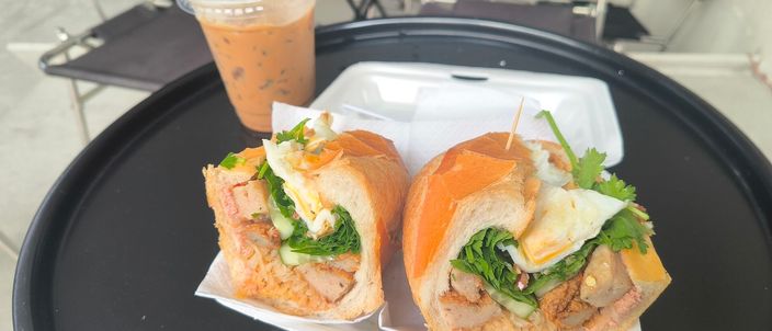 A bánh mì halved to show its thin crisp crust and airy crumb packed with meat, egg and coriander, in a foam takeaway box beside a glass of Vietnamese iced milk coffee on a Saigon café table.