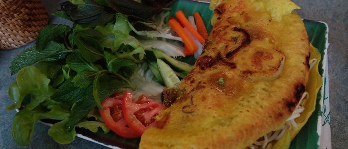 A folded golden, turmeric-yellow bánh xèo crêpe on a banana leaf, its filling of pork, prawn and bean sprouts showing at the open edge, served with a side of fresh lettuce, herbs, sliced tomato and pickled carrot.