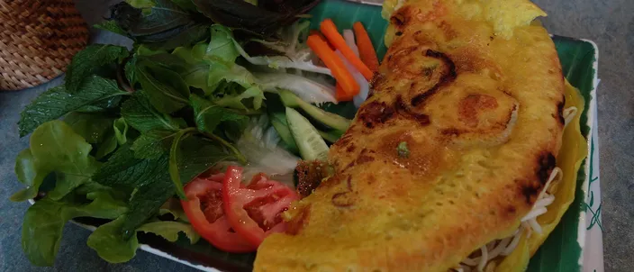 A folded golden, turmeric-yellow bánh xèo crêpe on a banana leaf, its filling of pork, prawn and bean sprouts showing at the open edge, served with a side of fresh lettuce, herbs, sliced tomato and pickled carrot.