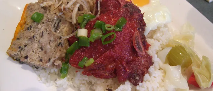 A plate of cơm tấm sườn bì chả: a mound of broken rice topped with a charcoal-grilled pork chop, a wedge of steamed pork-and-egg cake, a fried egg and chopped scallion, with pickled vegetables at the side.