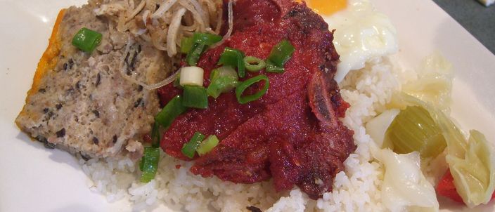 A plate of cơm tấm sườn bì chả: a mound of broken rice topped with a charcoal-grilled pork chop, a wedge of steamed pork-and-egg cake, a fried egg and chopped scallion, with pickled vegetables at the side.
