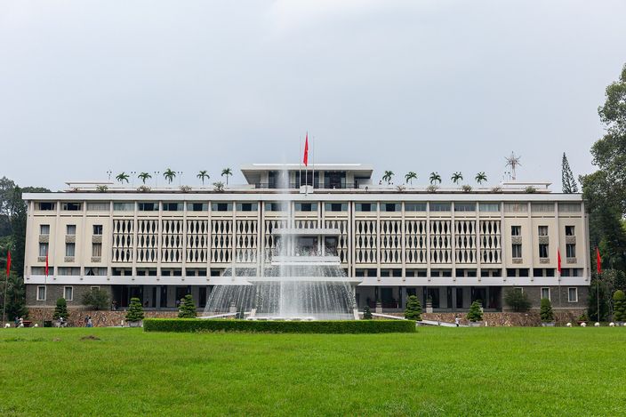 A wide four-storey modernist building faced with a lattice of pale concrete screens, seen head-on across a mown lawn, with a red flag on the roof, palms in planters along the parapet and a fountain playing in front of the entrance under an overcast sky.