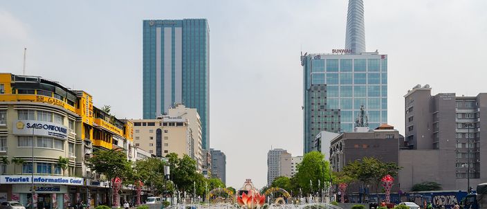A wide paved boulevard in bright daylight seen down its length, with a circular fountain and red flowerbeds in the foreground, low ochre colonial-era buildings and shopfronts along the left side, and glass office towers behind, including a tall tapering tower with a disc-shaped helipad at the right.