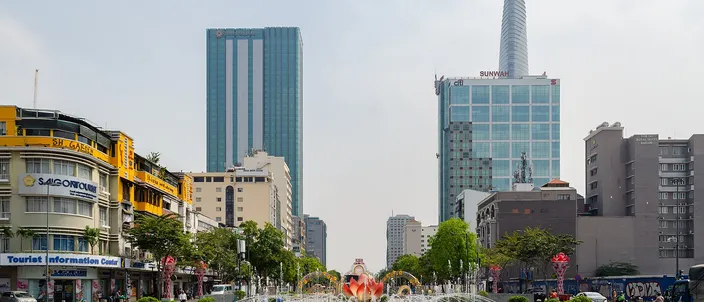 A wide paved boulevard in bright daylight seen down its length, with a circular fountain and red flowerbeds in the foreground, low ochre colonial-era buildings and shopfronts along the left side, and glass office towers behind, including a tall tapering tower with a disc-shaped helipad at the right.