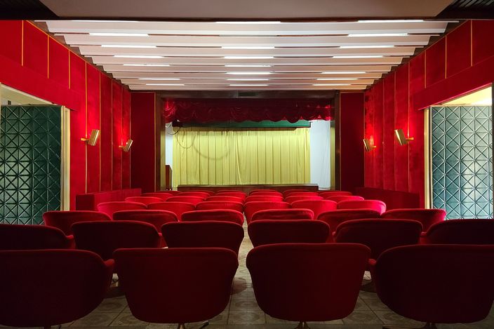 A small private cinema inside a 1960s presidential palace: five curved rows of deep red swivel armchairs facing a pale yellow stage curtain, red padded walls with brass sconces, a ceiling of long white light panels, and green patterned glass-block screens either side of the room.