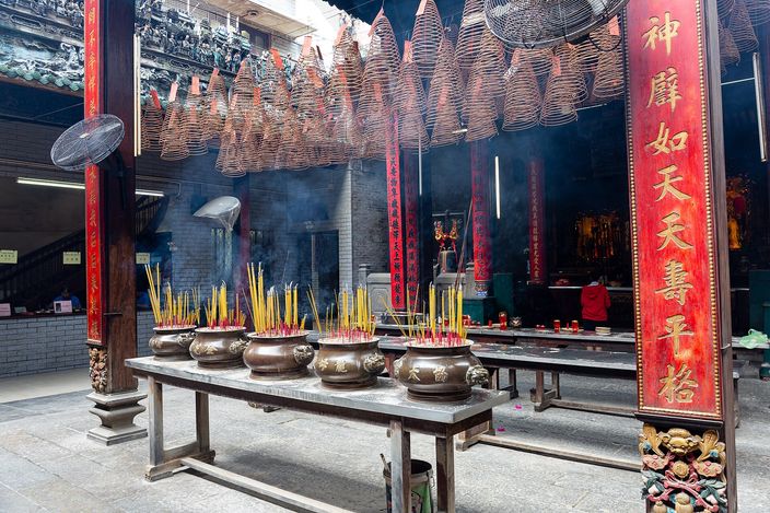 Rows of large conical incense coils hanging from the beams of a Chinese temple hall with smoke drifting up through them, bronze urns of burning incense sticks on a table below, and a red lacquered pillar carrying gilded Chinese characters at the right edge.