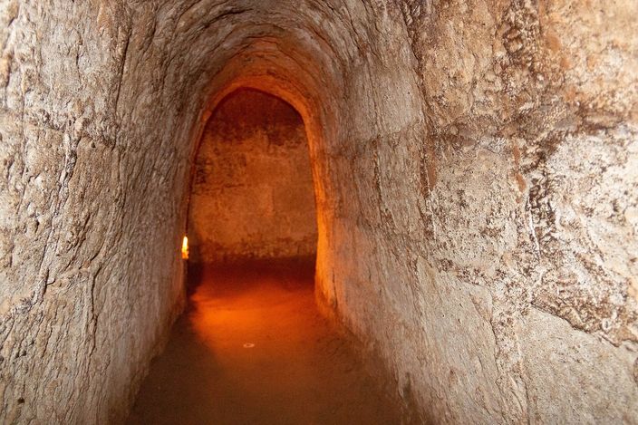 A lit underground tunnel of bare orange-brown earth, cut with a rounded arched roof and barely wider than one person, running back into a dim chamber.