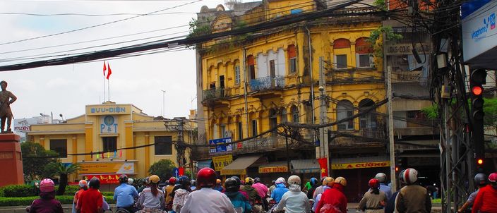 A crowd of motorbike riders stopped at a red light on a wide boulevard, with a weathered ochre French-colonial corner building of shuttered arched windows and iron balconies behind them, a low yellow post office flying a red flag to the left, and a tangle of overhead power lines across the top of the frame.