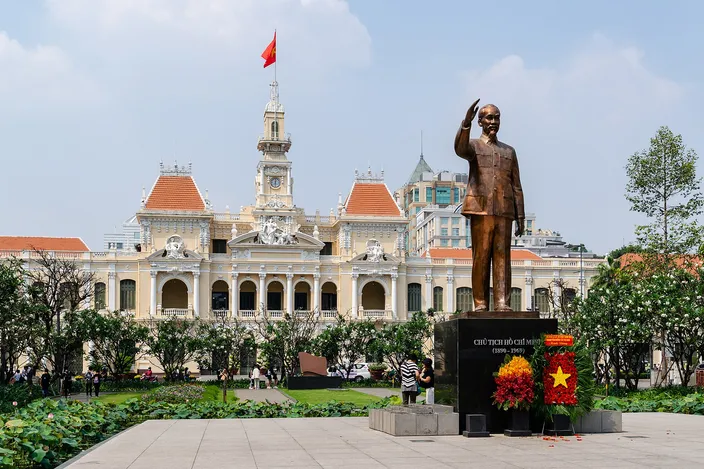 The golden bronze statue of Ho Chi Minh raising his hand, on a plinth with a wreath and Vietnamese flag, in front of the cream-and-white French-colonial People's Committee Building with its ornate clock tower, under a bright blue sky.