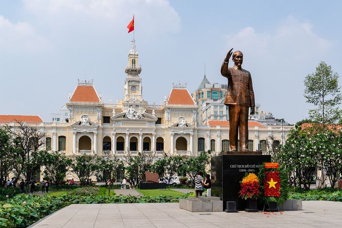 The golden bronze statue of Ho Chi Minh raising his hand, on a plinth with a wreath and Vietnamese flag, in front of the cream-and-white French-colonial People's Committee Building with its ornate clock tower, under a bright blue sky.