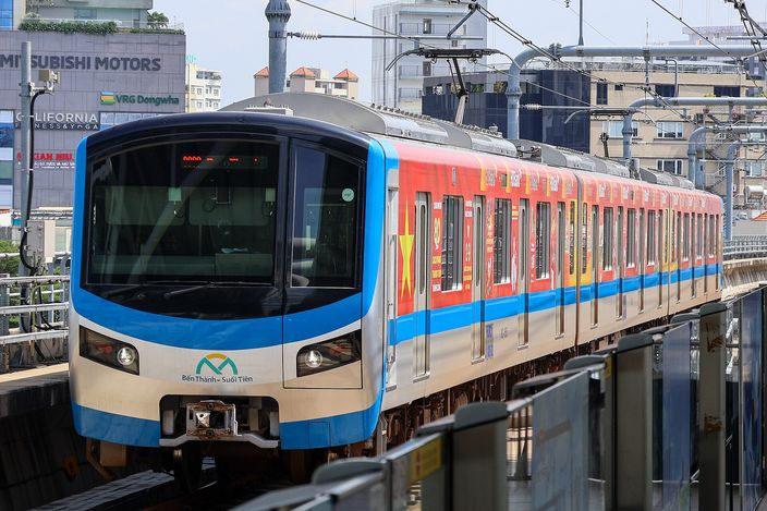 A blue, red and yellow Ho Chi Minh City Metro Line 1 train (marked "Bến Thành – Suối Tiên") arriving at an elevated station, with city buildings and Mitsubishi Motors signage behind under bright sun.