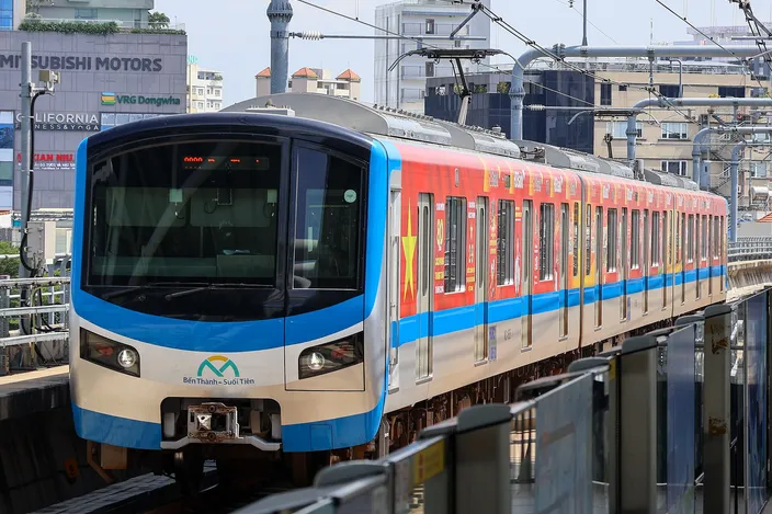 A blue, red and yellow Ho Chi Minh City Metro Line 1 train (marked "Bến Thành – Suối Tiên") arriving at an elevated station, with city buildings and Mitsubishi Motors signage behind under bright sun.