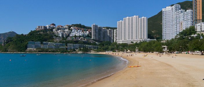 The wide golden crescent of Repulse Bay seen along the sand, with the line of shark-net floats visible across the turquoise water, residential towers stepped up the wooded hillside behind and green peaks closing the bay.