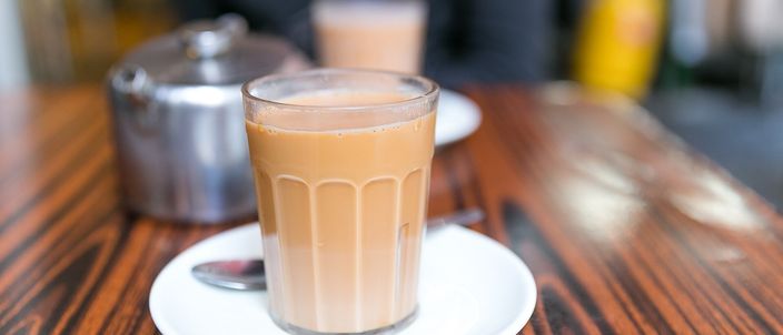A glass of Hong Kong milk tea, its milky red-brown colour showing the strong tea cut with evaporated milk, served on a saucer on a dark café table.