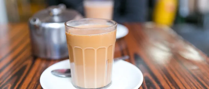 A glass of Hong Kong milk tea, its milky red-brown color showing the strong tea cut with evaporated milk, served on a saucer on a dark café table.