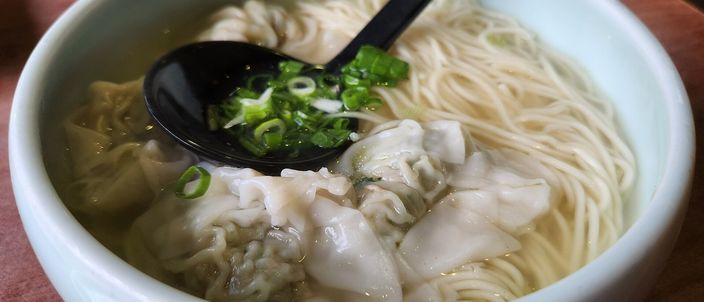 A bowl of Cantonese wonton noodle soup, a ceramic spoon lifting a prawn wonton from the clear broth over thin pale egg noodles and scallions.