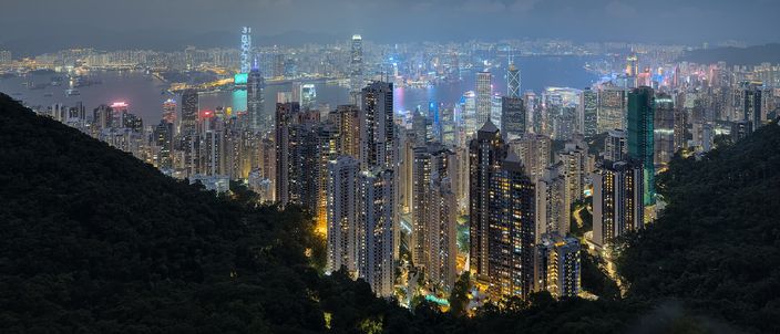 Dusk view from Victoria Peak looking down over the illuminated towers of Hong Kong Island packed against Victoria Harbour, with Kowloon beyond, wooded slopes framing the foreground