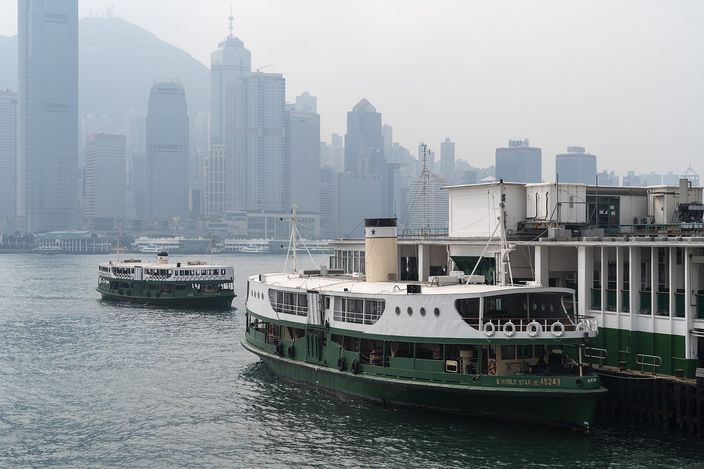 Two green-and-white Star Ferries at the Tsim Sha Tsui pier, one moored alongside and one crossing behind it, with the hazy towers of Hong Kong Island rising across the water