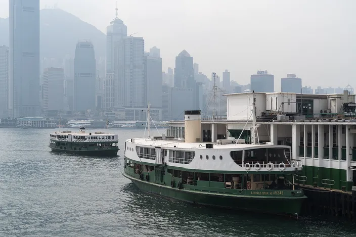 Two green-and-white Star Ferries at the Tsim Sha Tsui pier, one moored alongside and one crossing behind it, with the hazy towers of Hong Kong Island rising across the water