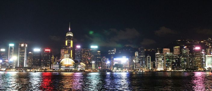 The lit towers of Hong Kong Island's north shore at night seen from across Victoria Harbour, their colours reflected in the dark water in the foreground